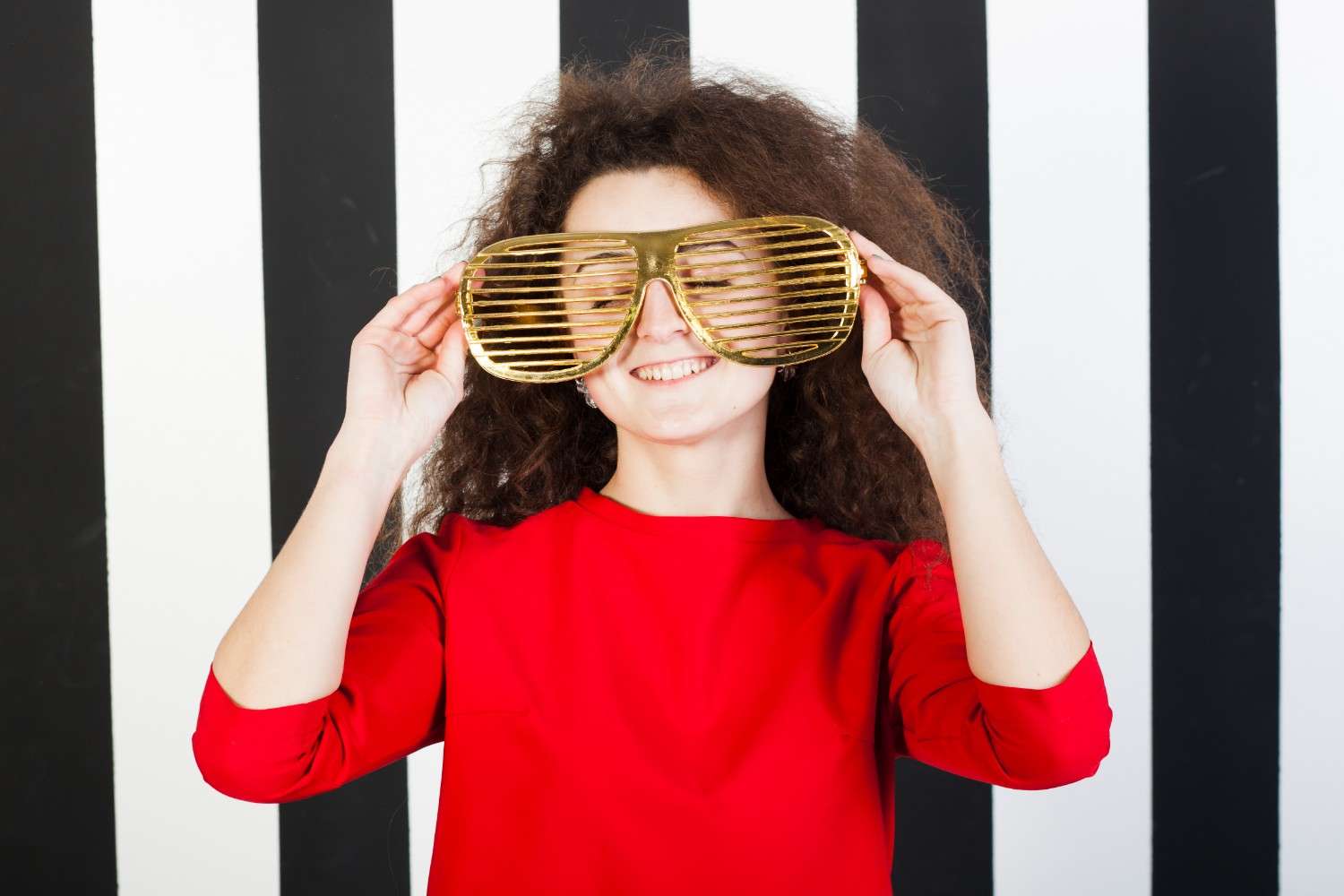 Portrait of a playful brunette girl with a bright smile, wearing a striped top, against a colorful striped background. The girl has her hands on her hips and is looking confidently at the camera.