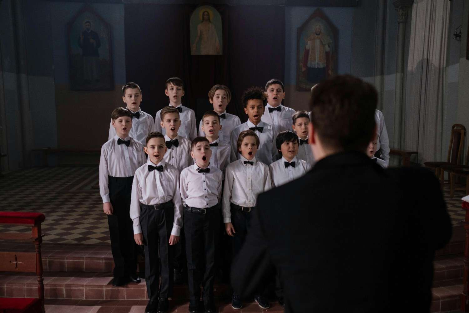 A group of children in a church setting, following a music conductor as they practice singing together.