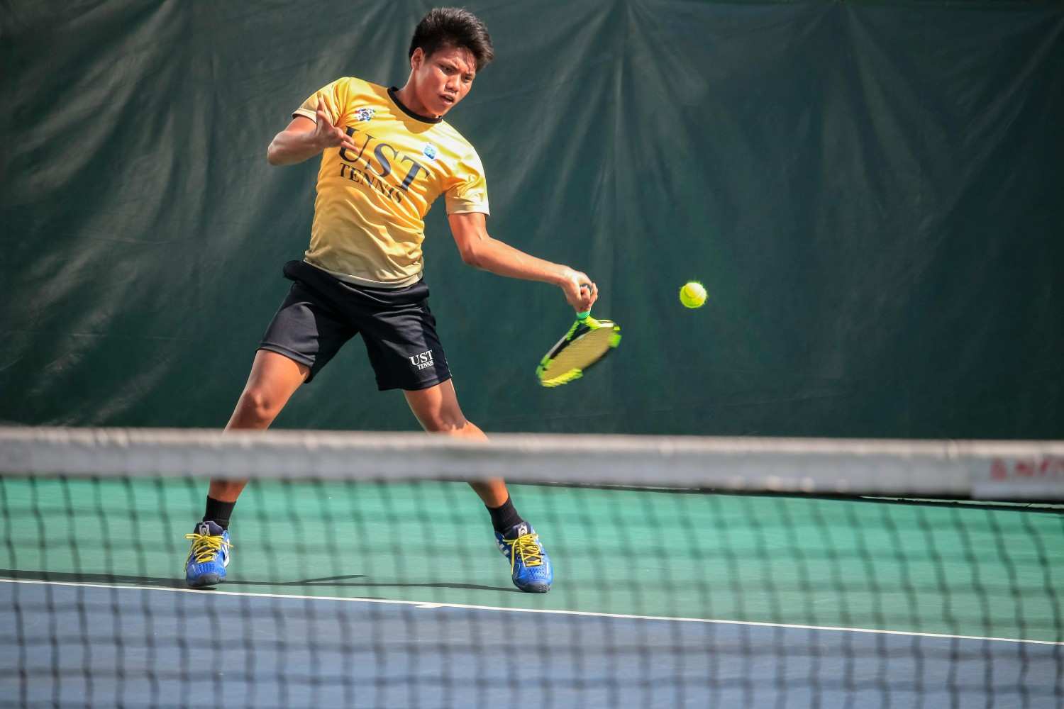 Man in mid-action hitting a tennis ball on an outdoor court, wearing a white shirt and black shorts, with a bright blue sky and green court in the background.