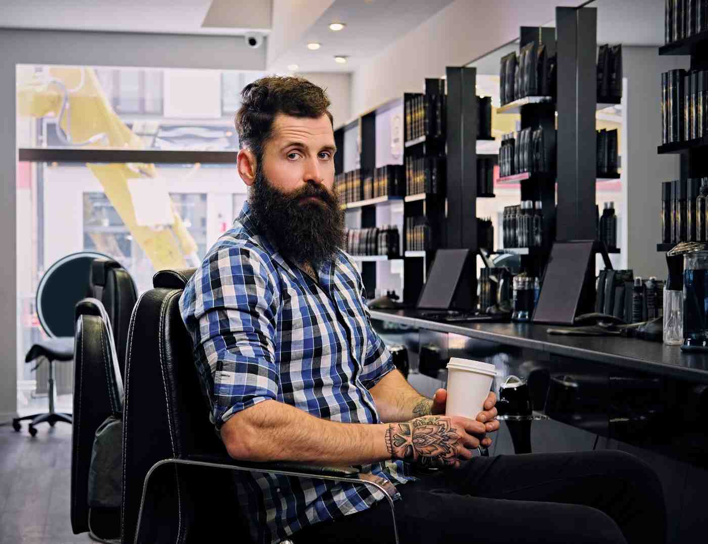 A stylish bearded man in a fleece shirt drinks coffee while sitting in a hair salon, looking relaxed and enjoying his time